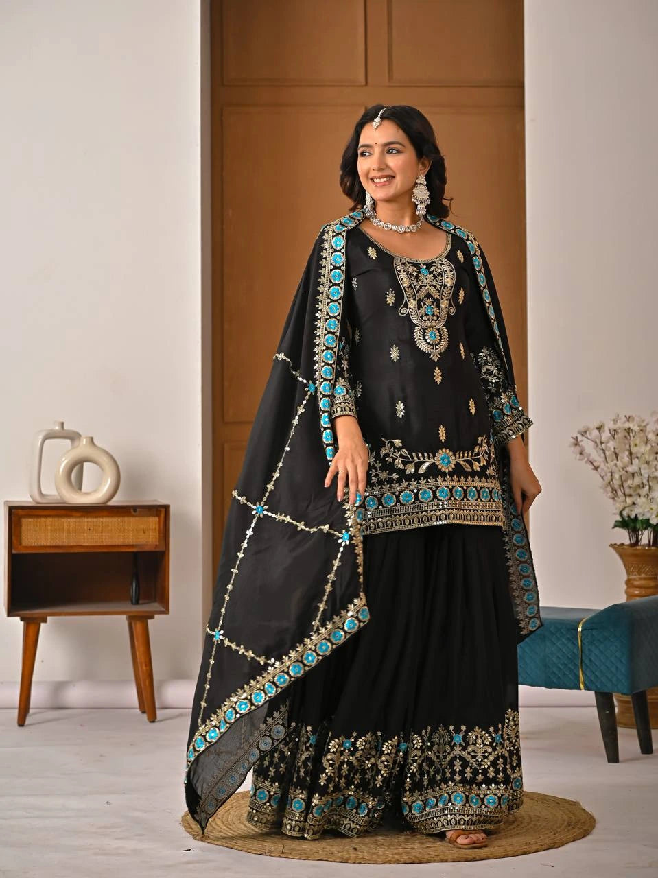 Woman in a black embroidered traditional outfit with turquoise details in a room setting.